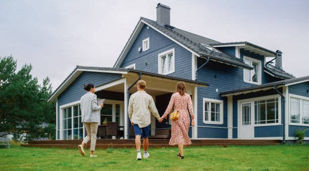 Young Couple Viewing Property for Sale, Talking with Professional Real Estate Agent Outside the House on a Summer Day. Young Beautiful Family are Ready to Become New Homeowners.